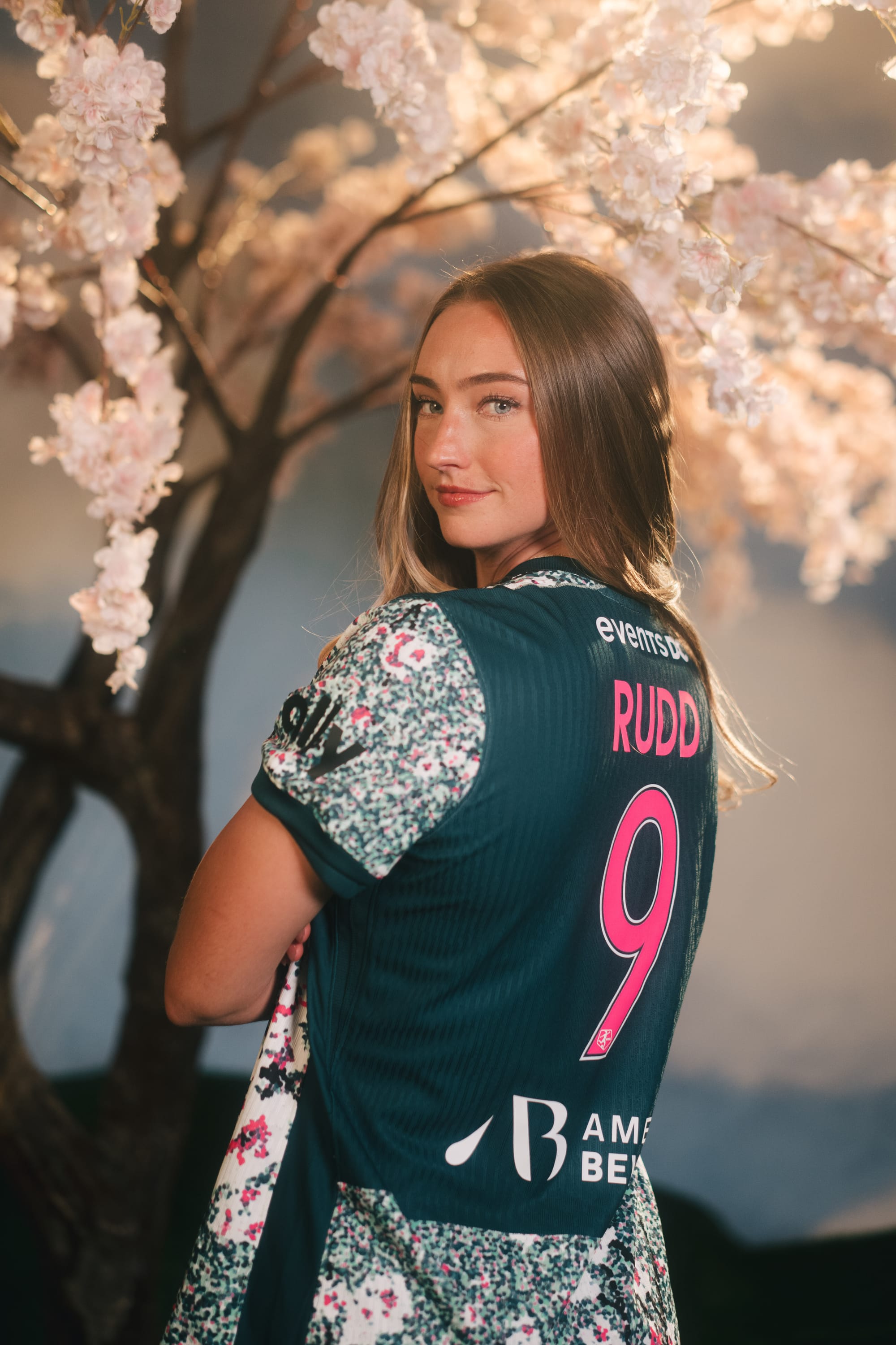 A photo of Tara Rudd wearing the new "Spirit in Bloom" kit. She is facing away but has turned to her left to look back at the viewer, which shows the new kit's vibrant pattern on the sleeve and lower back, as well as the solid dark green section on the middle and upper back.