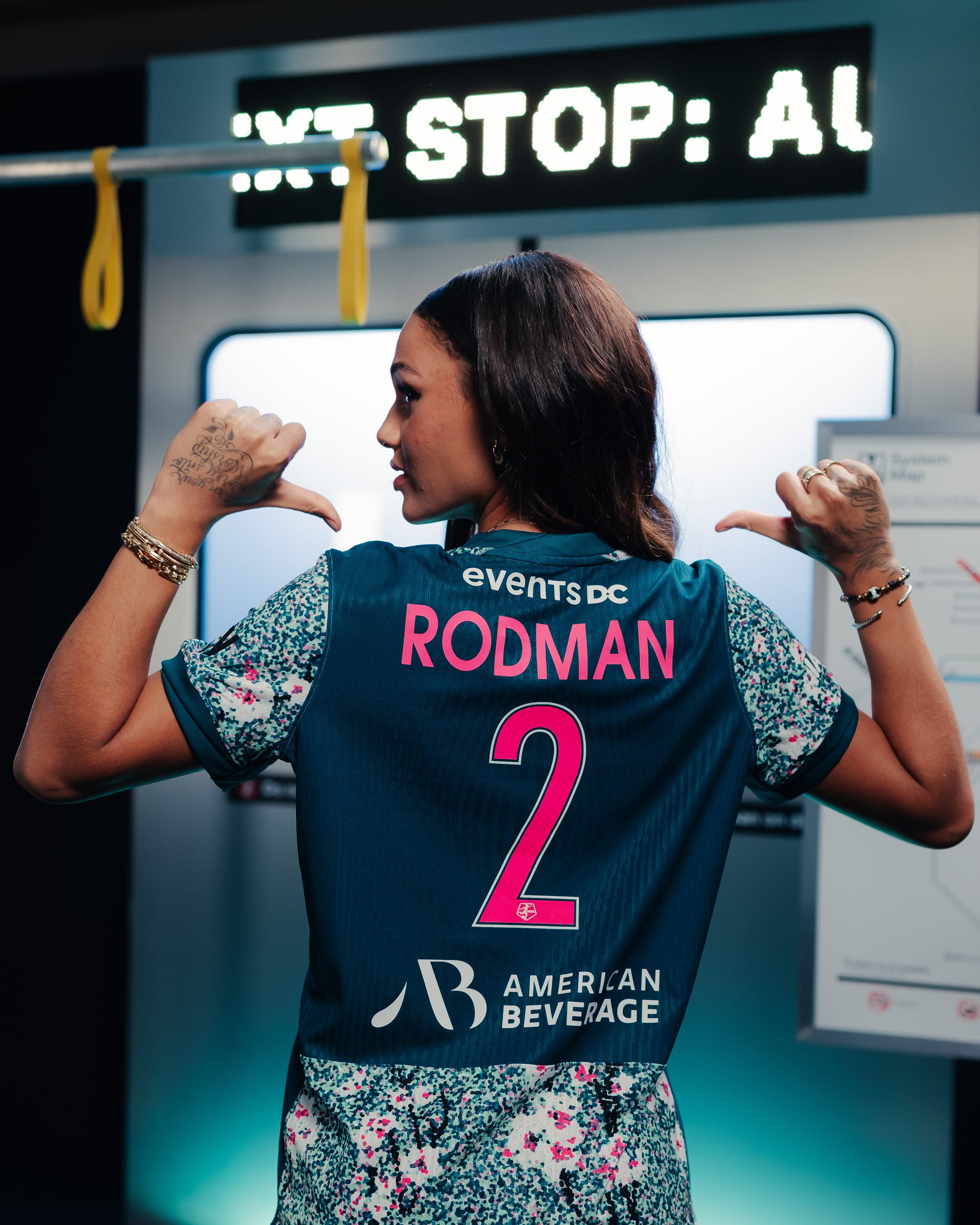 A photo of Trinity Rodman wearing the new "Spirit in Bloom" kit. Rodman has her back turned to the viewer, and is gesturing with both thumbs at the name on her kit. Her name and number are in bright pink letters amid a solid patch of dark green. The sleeves and lower back of the kit appear like the front, with dark green and white speckles alongside cherry blossoms and branches.